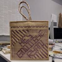 Item 1, side 1. A basket made from finely woven material in natural color and purple dyed fibers with square sides. The purple fibers are woven in a pattern that spells out "HEY GOODBYE RYAN" on one side.